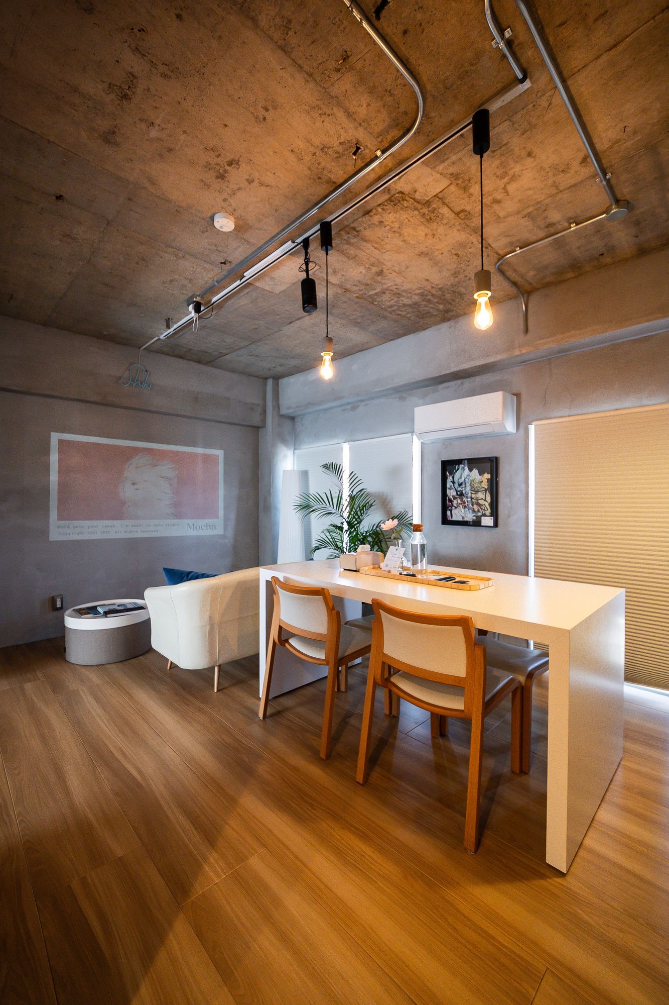 Pale dining table and sofa on an oak floor beneath a bare concrete ceiling, artwork projected on the wall