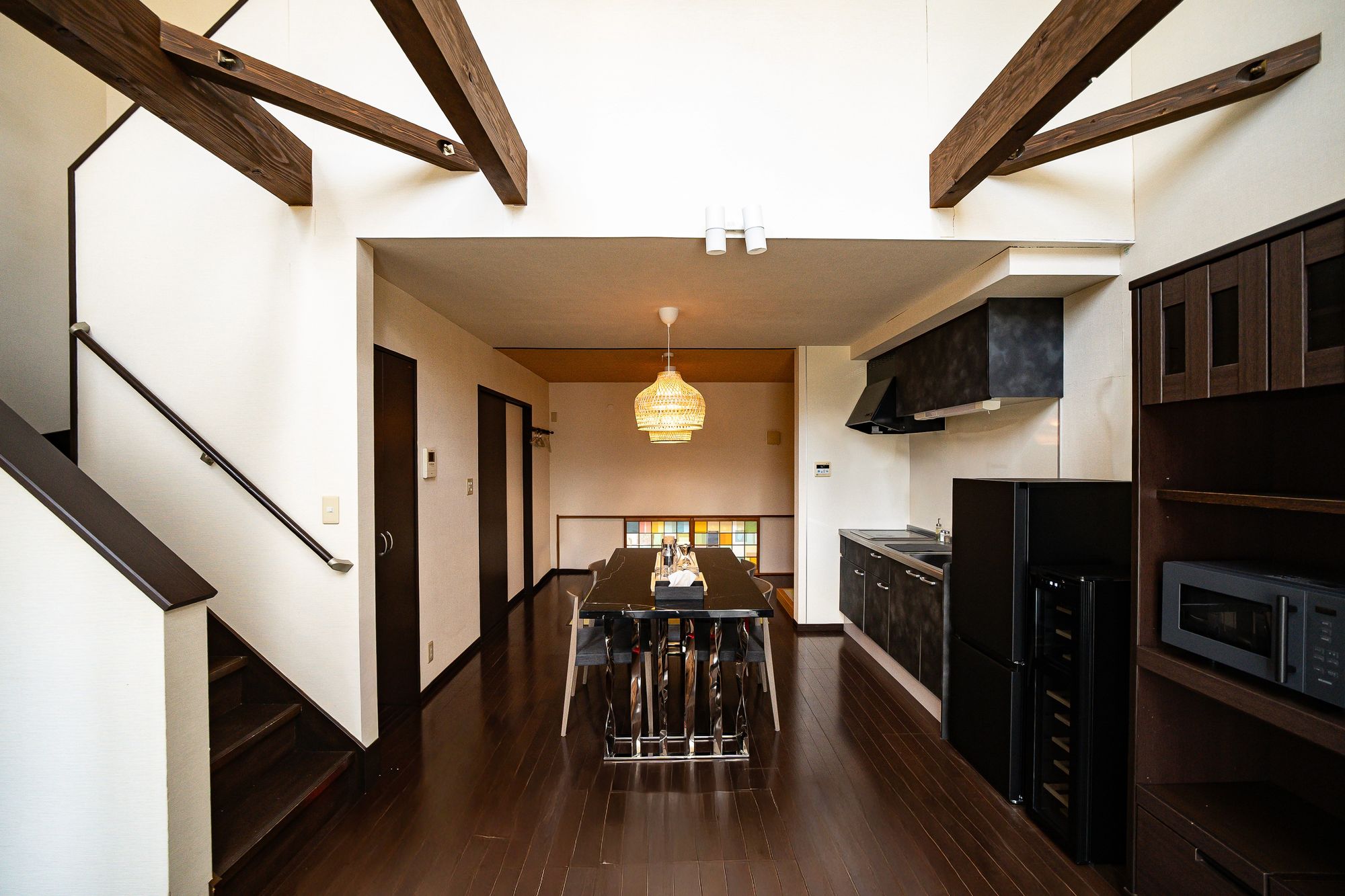 Open-plan kitchen and long dining table under exposed timber beams, stair rising to the left